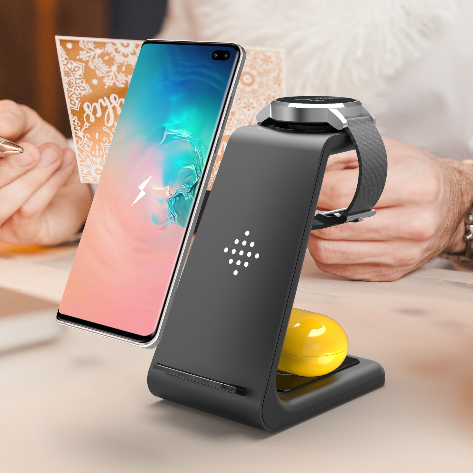 Black multi-device charging stand with smartphone, smartwatch, and yellow remote control on a blurred background.